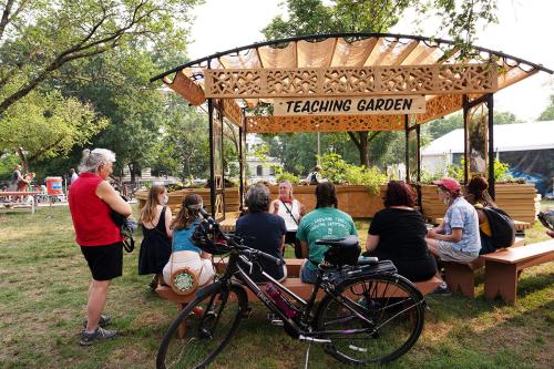 Under a wooden structure with a sign that reads Teaching Garden, a woman speaks with a small group of people seated on benches.