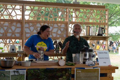 A woman laughs while grating taro over a cutting board, while a man gestures with his hand, mid-speech. 