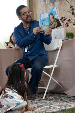 A man, seated, holds up an illustrated picture book, reading from it. A young child seated on the ground looks up at the book.