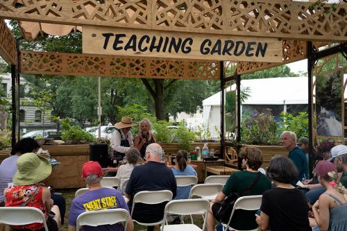 Under a wooden lattice structure and hand-painted sign that reads Teaching Garden, two women present for a small crowd seated in folding chairs. Around them, raised planter beds are filled with green plants.