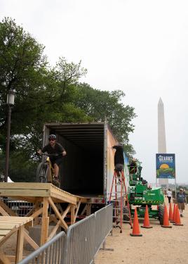 A person rides a bike out of a semi-truck trailer onto a wooden boardwalk while two people on lifts and ladders spray paint the side of the trailer. In the distance, a banner for the Ozarks program and the Washington Monument. 