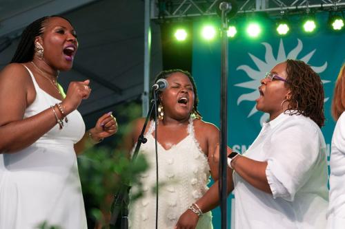 Three women in white dresses sing on stage under green lights.
