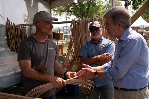 Three men talk outdoors. One holds a handful of flat strips of wood, with a tool tucked under his arm.