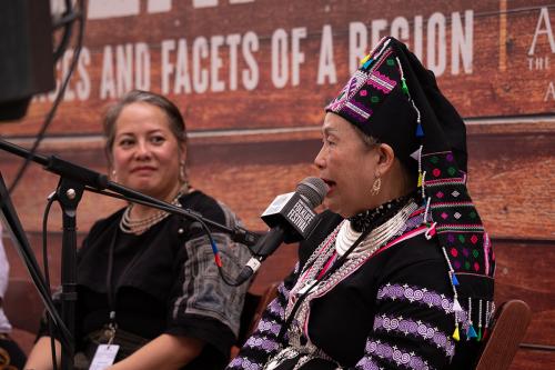Two Asian women sit on stage, as the older speaks into a Folklife Festival microphone. She wears elaborate silver jewelry and black top and headdress with colorful embroidery.