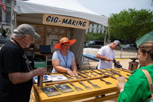Four people stand around an outdoor table set with books and metal pronged spears. Above them, a hand-painted sign reads Gig-Making.