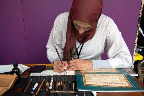 A woman in a maroon  hijab and white shirt leans over a desk, writing calligraphy. In front of her are a calligraphy book, a cell phone, and a black case with several pens.