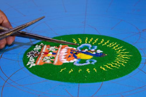 In colored sand, a blue figure at the center of a circular design on a blue table. 
