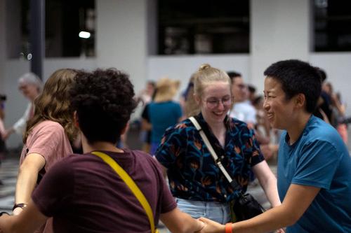 Four people dance in a square, hands joined and smiling. A crowd behind them dances as well.