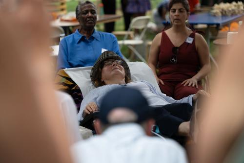 Framed by bare arms out of focus on either side, a person lays in a reclining chair, eyes closed, while those seated around them sing.