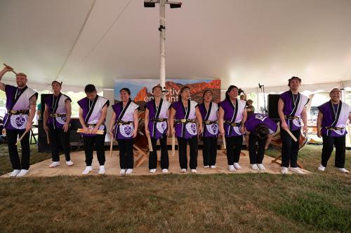 Eleven people in matching purple and white tops and black pants, each holding a pair of wooden drum beaters, line up to take a bow under a white tent.