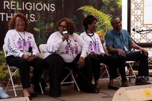 Three women and a man sit in a row on an outdoor stage, all laughing. The three women wear matching white, long-sleeves shirts with a photo of themselves on it.