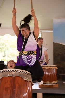 Mid-air, a woman jumps while playing a wooden barrel drum, both sticks raised above her head.