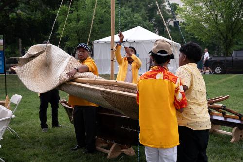 Four  men work to raise the sail on an outrigger canoe in the grass.