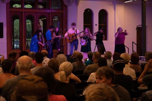On stage, a five-piece string band perform in front of a crowd. Also on stage are a videographer and an ASL interpreter.