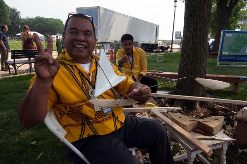 Sitting under a tree, two men in yellow button-up shirts give a peace sign and thumbs up to the camera. The man in front holds a small, simple scale model of a boat.