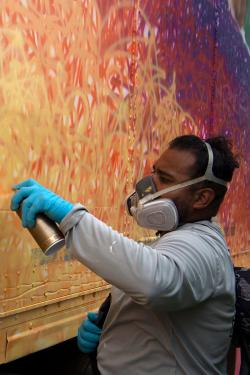 A man in a gray shirt and hazmat mask spray paints a trailer with various circles of red, orange, yellow, and purple. 
