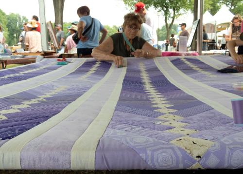 A purple patchwork quilt with different stripes and diamonds with a woman threading a needle through the quilt at the far end away from the camera.