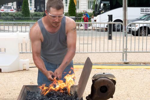 A man in a tank top and safety goggles holds a piece of metal over flames in a forge.