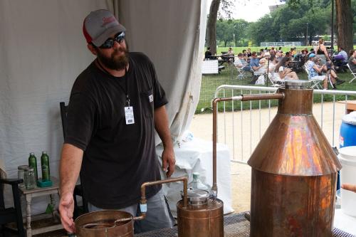A man with short beard and sunglasses gestures toward one of the copper pots of a still. In the background, a crowd sits in folding chairs.