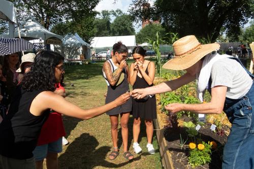  A woman in a hat bends over to hand a small plant to a visitor, with two visitors smelling plants in the background.
