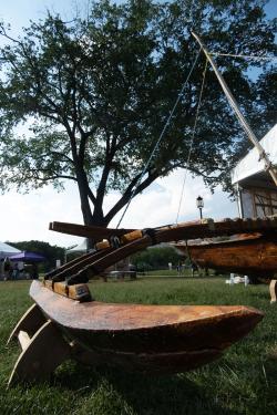 From ground level, a wooden outrigger canoe with mast pole stretching toward blue sky.