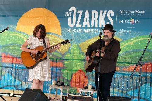 A woman and man play acoustic guitars on stage, in front of a colorful backdrop of a mountain scene and the program title, The Ozarks. 