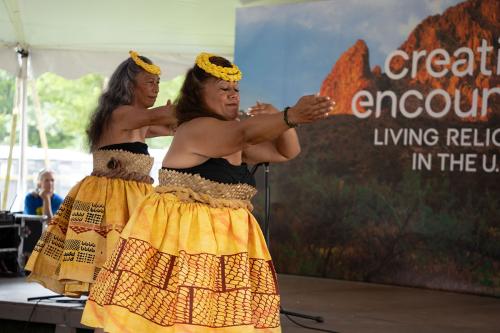 Two women in yellow skirts with woven waistbands and black tops thrust their right arms out straight ahead, their left arms tucked in near their cheeks.