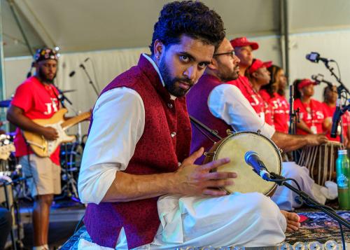 A man holding a drum in a red vest stares straight at the camera, while other qawwali singers and gospel singers perform on the stage.