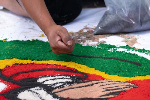 Close up on the hands of a Guate-Maya D.C. artist, pouring black dyed sawdust to outline La Virgen de Guadalupe’s shawl in the second alfombra.