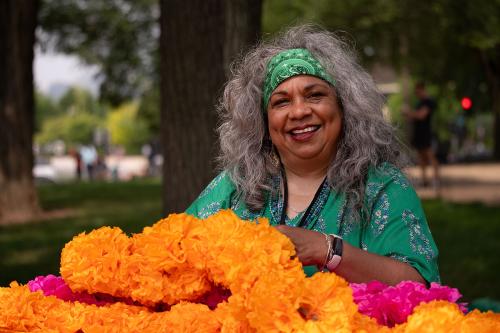 A woman with a green headwrap and green blouse smiles over a pile of yellow and pink marigolds.  