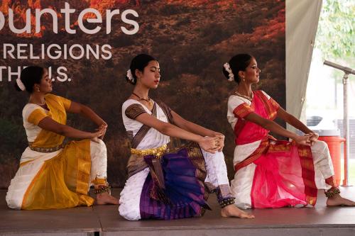 Three dancers kneel on stage with matching white tops with colorful sashes.