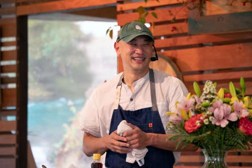 Tim, the chef, stands behind the demonstration kitchen counter, clutching a white dishrag and laughing.