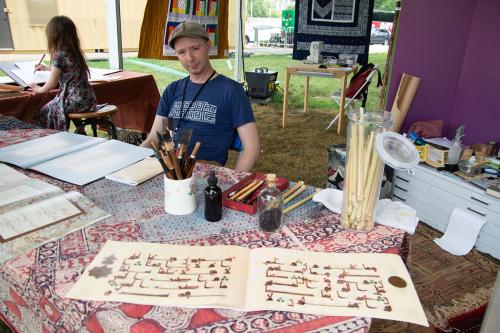 A man sits at a desk outdoors, surrounded by calligraphy tools and books. A young child behind him practices calligraphy.