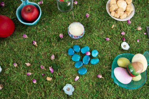 From above, a tabletop display with green turf, smooth turquoise stones, pink dried flower petals, a red apple in a bowl, painted eggs in a dish, and other small items.