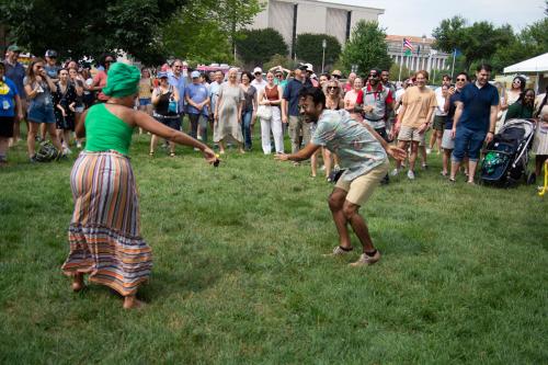 A woman in a green head wrap and top with striped pants dances with a man in a Hawaiian shirt and khakhi shorts. A crowd of visitors watch on in excitement, with the American History Museum and the flag of Puerto Rico visible in the background.