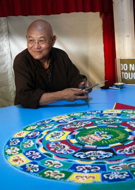 A man smiles peacefully, sitting in front of a colorful sand mandala.
