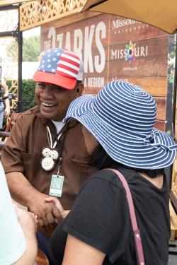 A man in a hat with the American flag smiles and shakes the hand of a woman in a blue and white striped hat. 