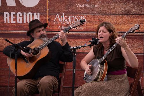 A man plays a guitar on the left, looking toward the tuning pegs, while a woman closes her eyes and sings into a microphone, playing the banjo.