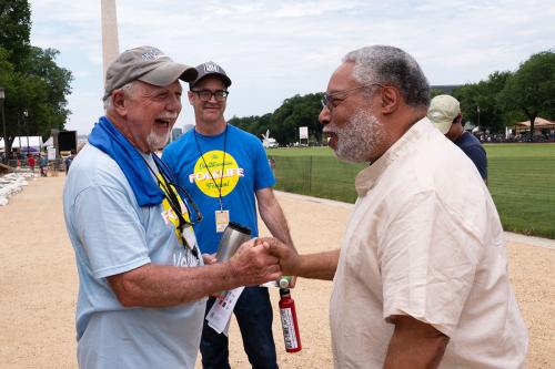 Two elder men joyfully shake hands on the National Mall, with the Washington Monument in the background.