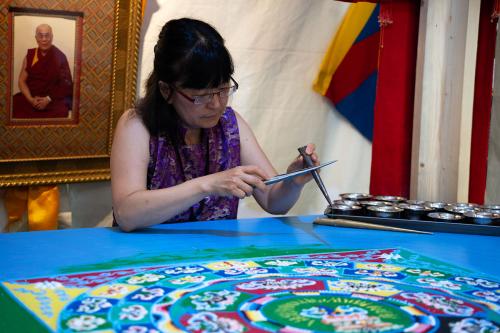 A woman works with two thin metal tools over a colorful sand mandala. Behind her, a portrait of the Dalai Lama. 