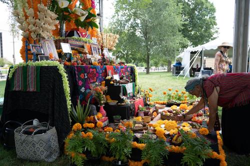 A woman bends down over an outdoor altar, adjusting orange paper marigolds. The tiered altar is set with plants, food, picture frames, and other objects.