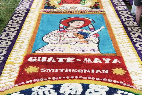 Detail of the second alfombra featuring a woman wearing traditional white huipiles of Guatemalan cofradías onto of a light blue sawdust background, with a Guate-Maya and Smithsonian logo below made out of rice.