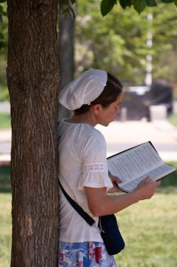 A woman in a white head wrap singing with an open hymn book in hand, leaning against a tree.