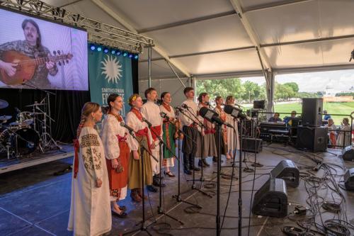 Nine singers in traditional Ukrainian dress sing into microphones on the Ralph Rinzler Main Stage, with a video of a man playing a traditional Ukrainian plucked string instrument.