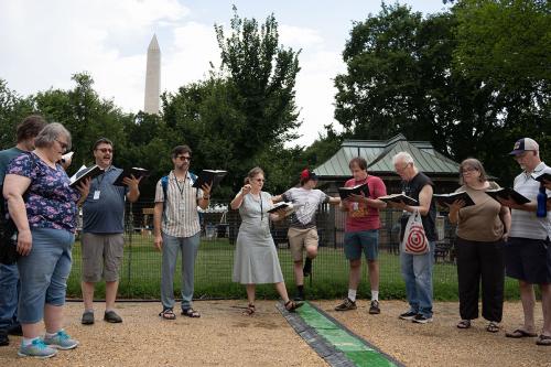 Ten people form a horseshoe shape, each holding and singing from a hymnal. There are puddles on the gravel ground, and the Washington Monument is in the background.