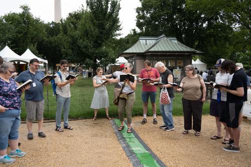 A woman leads a group of people standing in a circle standing on gravel in front of the National Mall, the Washington Monument visible in the background.