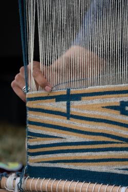 Close-up on a person weaving on an upright loom, using white, blue, and yellow yarn to created a zig-zag pattern.