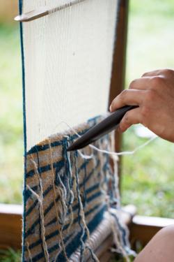 Close-up on a person weaving on an upright loom, using a wooden tool to pass threads through.