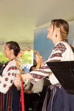 A woman smiles in profile while shaking a tambourine while another woman smiles facing the left, both dressed in traditional Ukrainian dress.