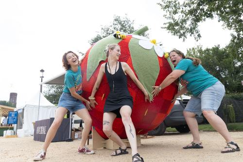 Three women pretend to be holding a giant crochet strawberry back from rolling away, wide expressions on their faces.